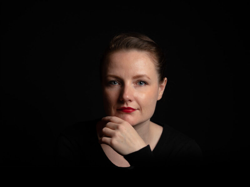 A woman whose face and hand are lit, but the rest of the image is dark. The hand rests gently across the chin.