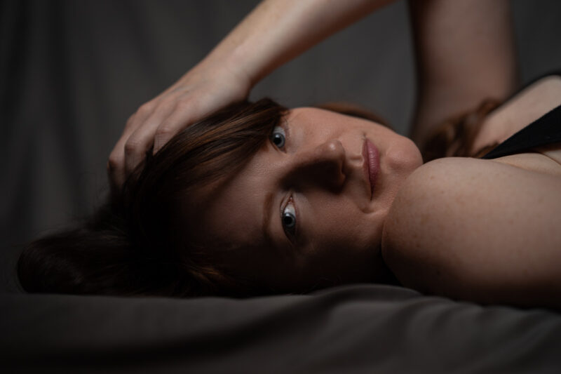 A women is lying on a medium grey background on the floor looking at the camera with her head turned to the right. Her left hand rest softly on the left side of her head in her hair.