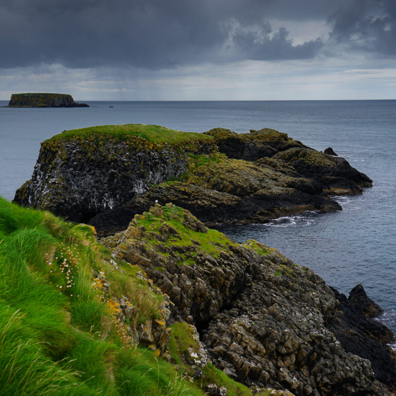 A storm brews off the coast of Northern Ireland. there are bright green grasses on dark, cracky cliffs.
