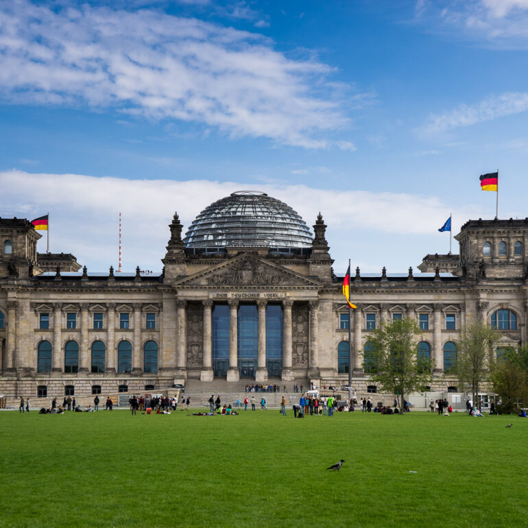 The Reichstag Building in Berlin. The sky is blue with some white clouds. The foreground is bright green grass