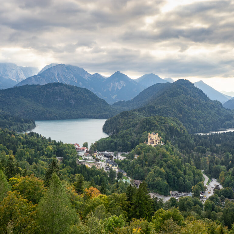 Schloss Hohenschwangau as seen from Schloss Neuschwanstein in Germany. The sky is filled with fluffy white clouds that hide the sun, but the rays are poking through. There are mountains and trees surrounding the castle and nearby town.
