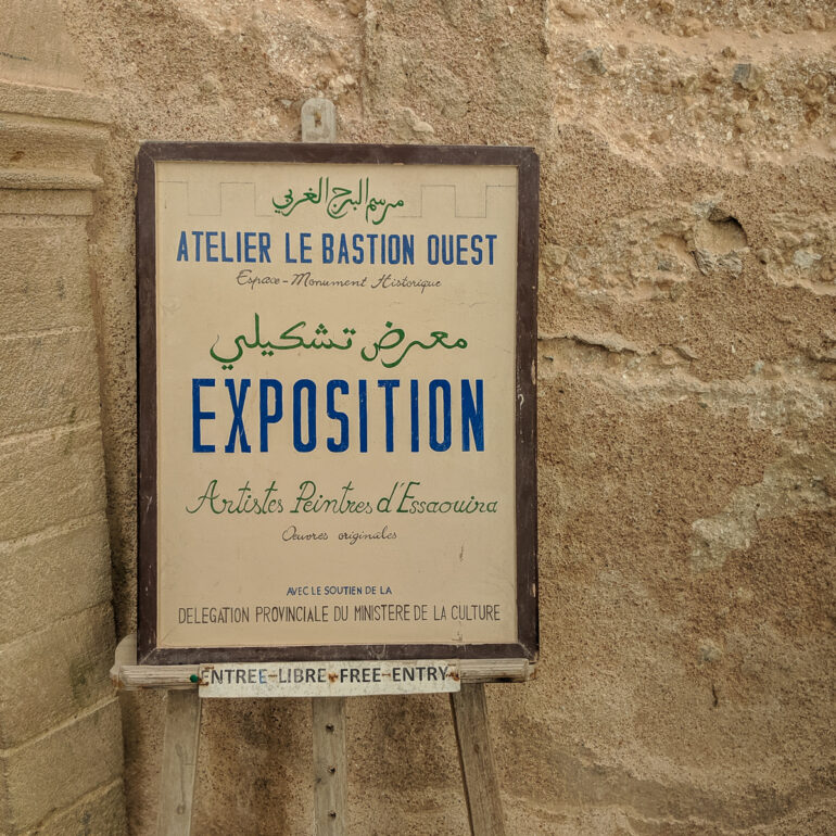 A sign is on a stand against a wall. The sign is for an art exhibit and is written in Arabic and French