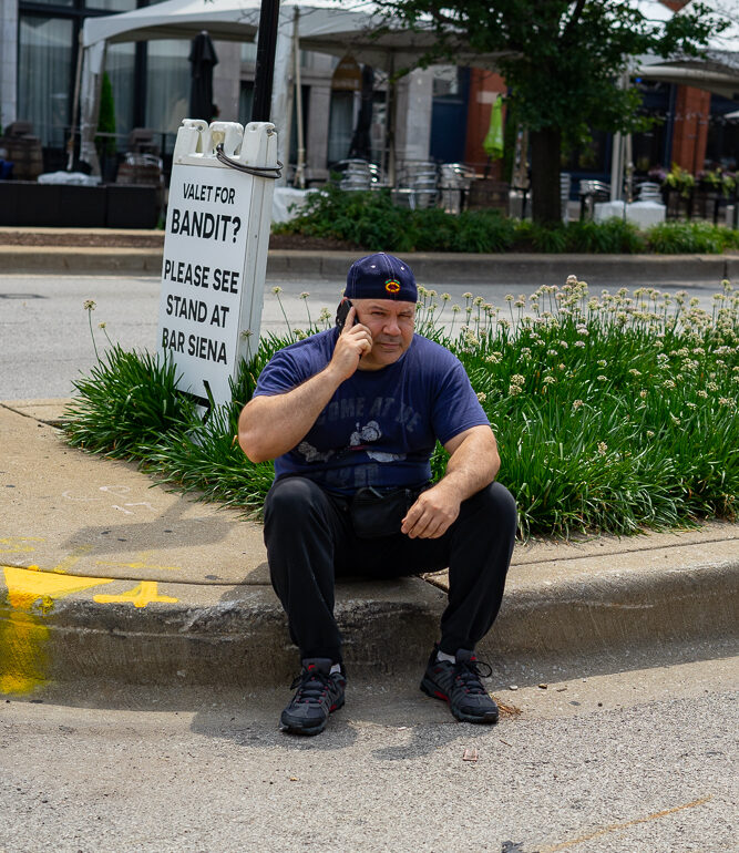 A man sits on a curb of the street talking on a cell phone.