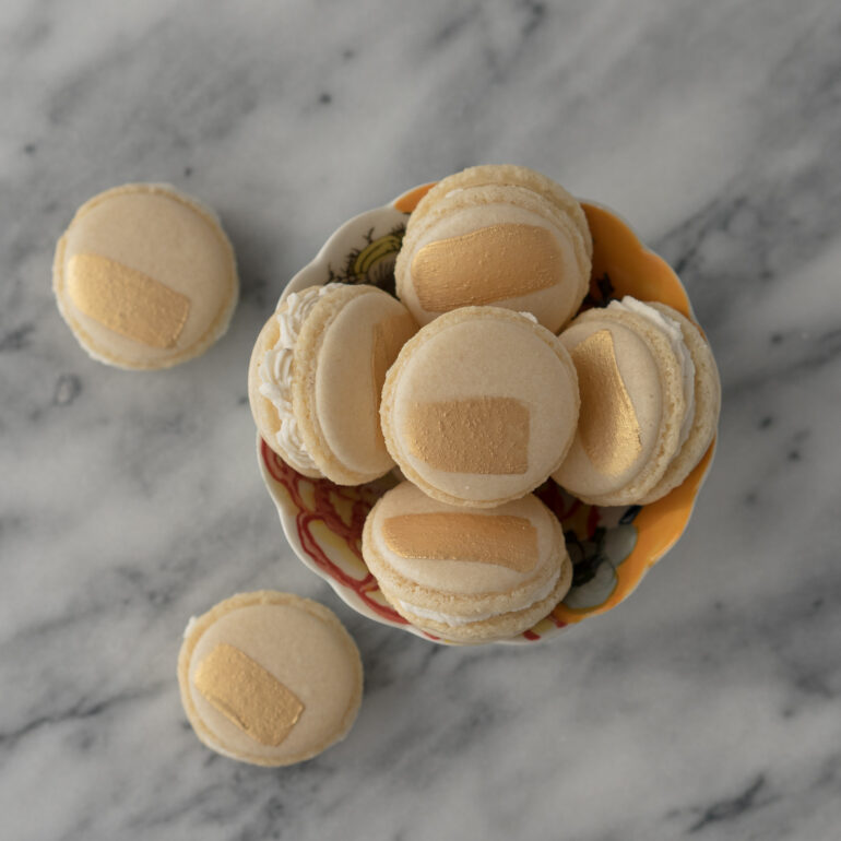 A small tea cup is filled with vegan vanilla macarons that have been painted with a stripe of gold edible paint.