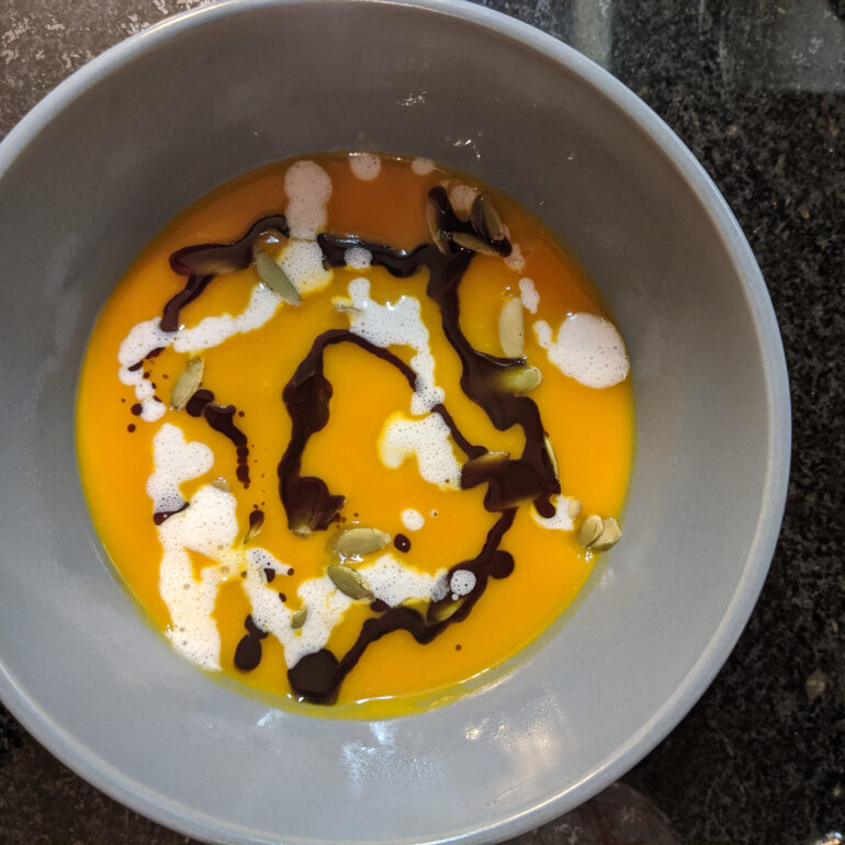 A bowl of pumpkin soup is drizzled with pumpkin seed oil and cream. Pumpkin seeds are scattered on top.