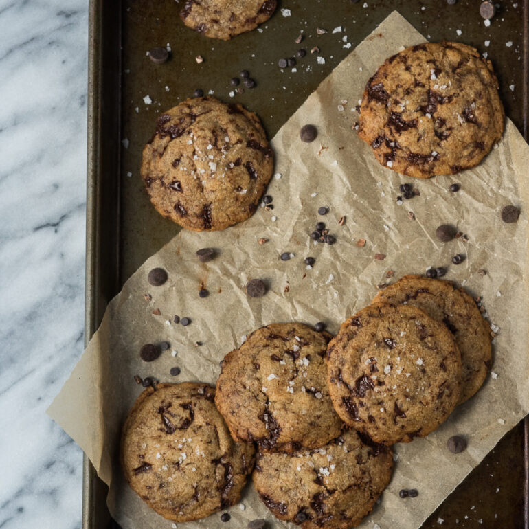 Chocolate chip cookies are littered on a baking sheet with scatter pieces of chocolate around the cookies.
