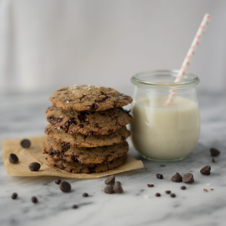 A stack of chocolate chip cookies is next to a cup of milk with a white straw and pink hearts. Chocolate chips are scattered around them.