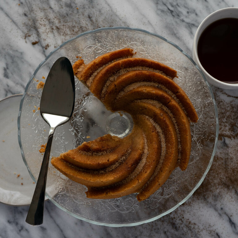 A bundt cake is on a clear cake stand with a wedge of the cake cut out of the cake. There is a serving tool & a plate to the left and a cup of chai tea to the right.