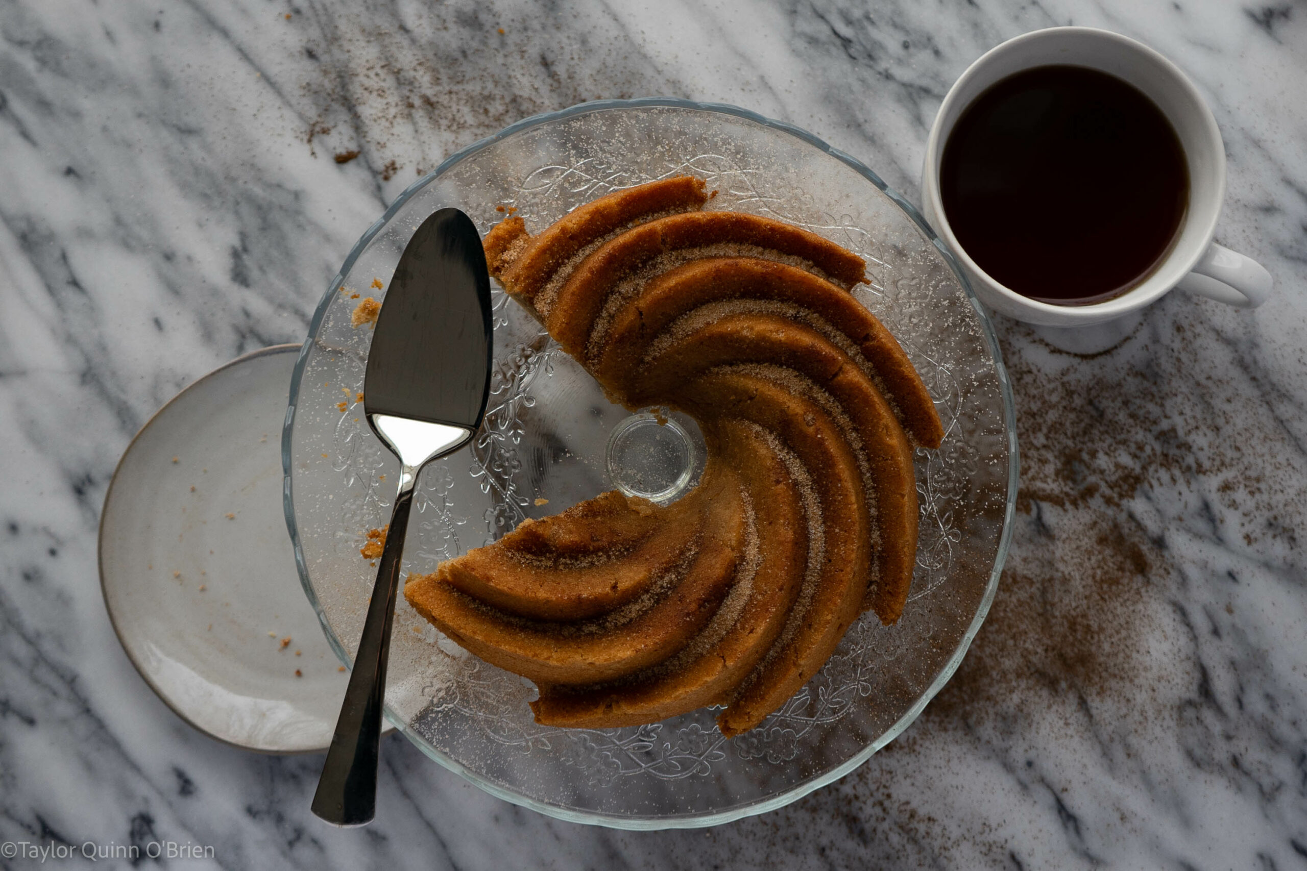 A bundt cake is on a clear cake stand with a wedge of the cake cut out of the cake. There is a serving tool & a plate to the left and a cup of chai tea to the right.