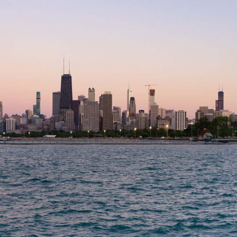 The Chicago skyline at sunset. The sky is a soft shade of pink and purple with Lake Michigan in the foreground.